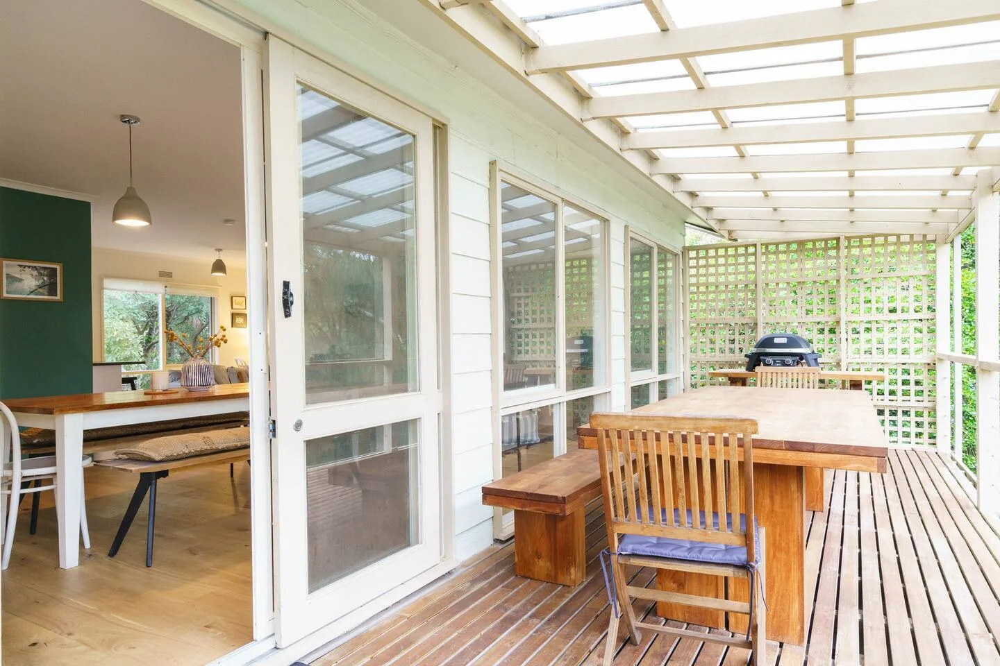 Una terraza de madera cubierta tiene una mesa de comedor y asientos junto a grandes ventanas.
