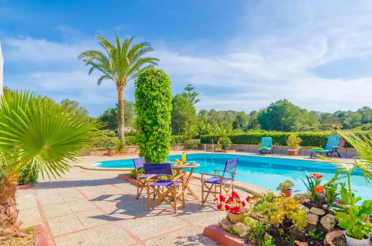 An outdoor swimming pool is surrounded by patio seating and tropical plants.