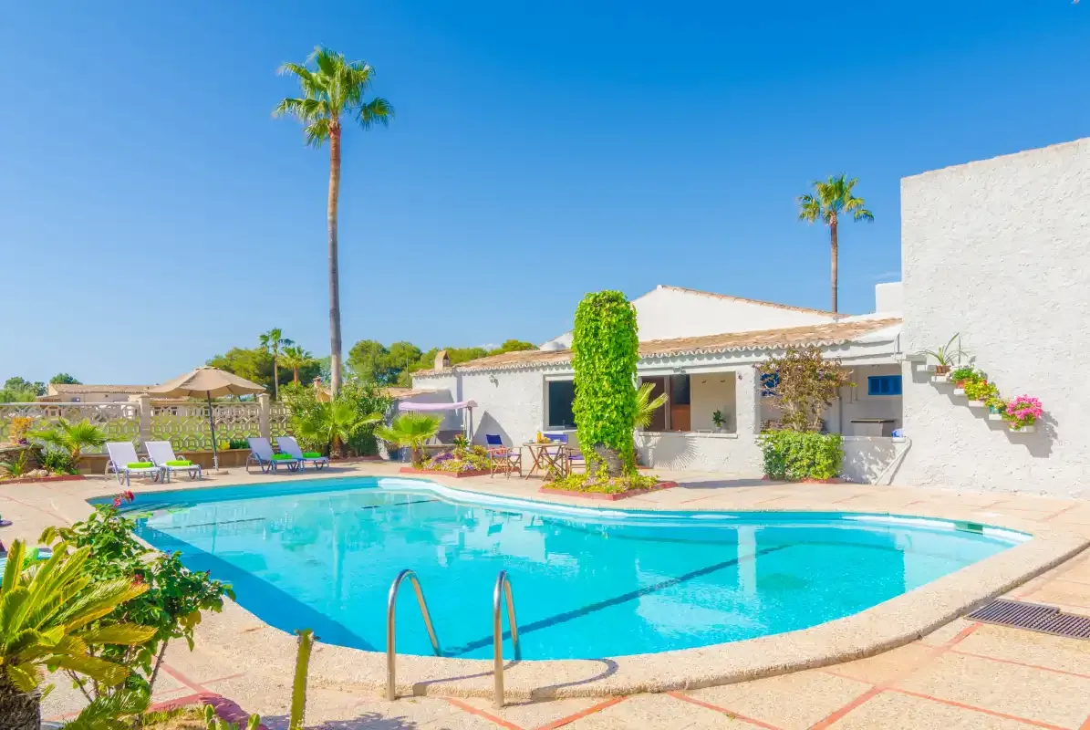 A swimming pool sits beside a low white building and surrounding palm trees.