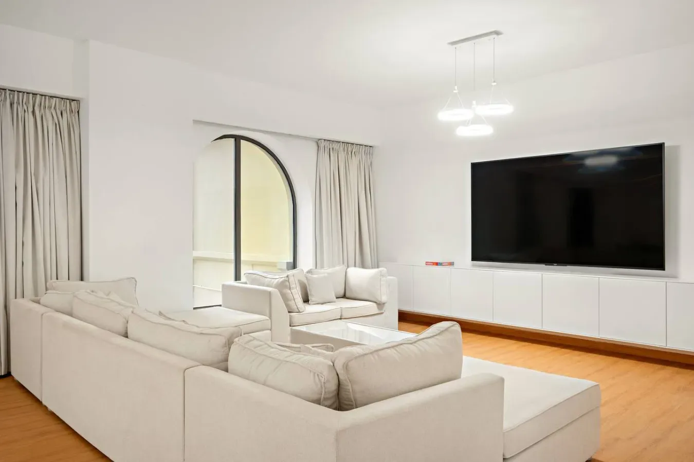 Una sala luminosa con asientos color crema, un televisor grande y ventanas con cortinas.