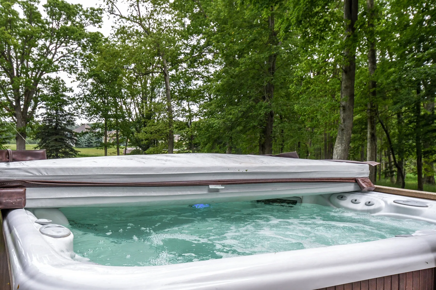 An outdoor hot tub sits beneath leafy trees, with its cover partially open.