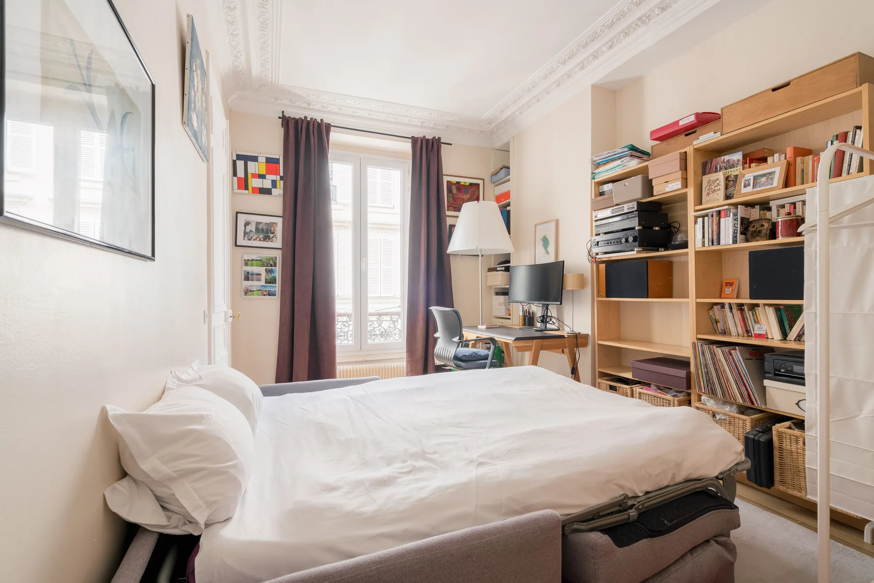 A bright bedroom with a bed, tall bookcases, and a desk beside the windows.