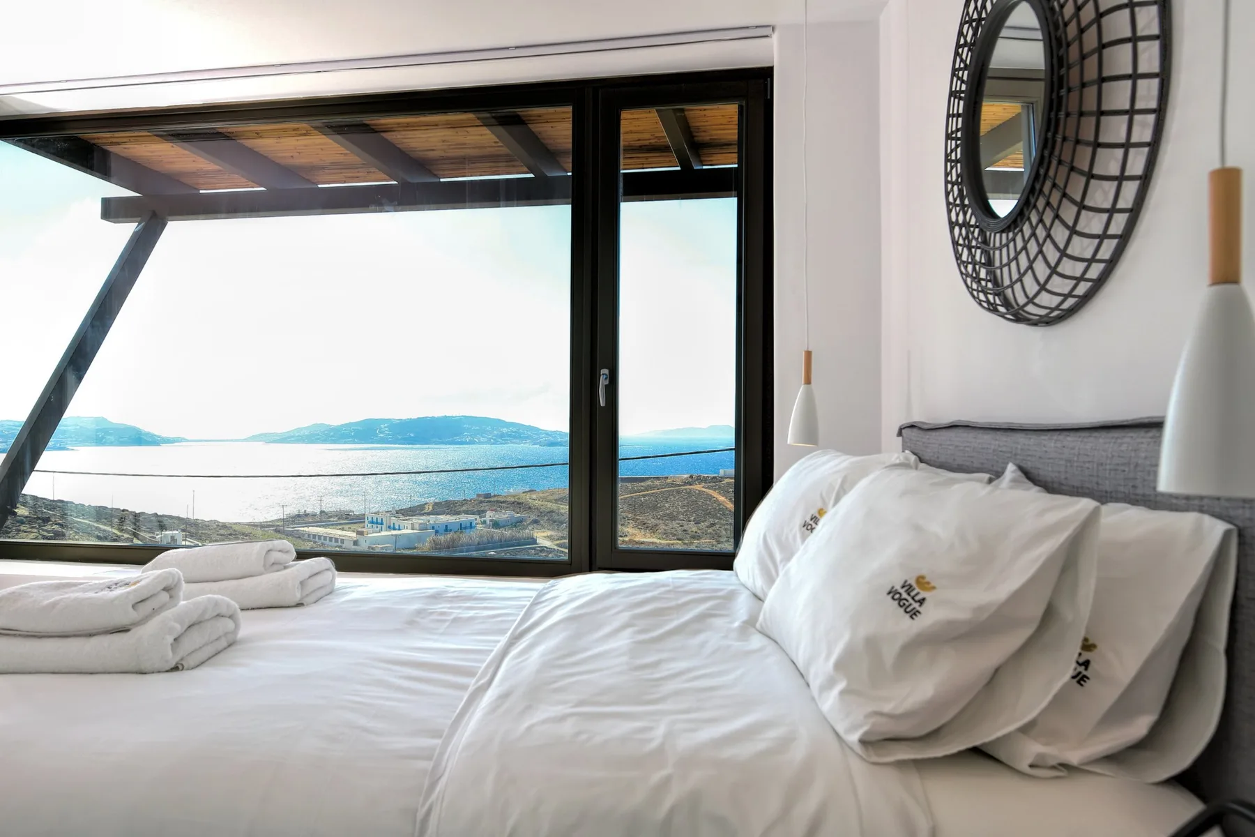 A bright bedroom with white bedding and a wide window overlooking the water.