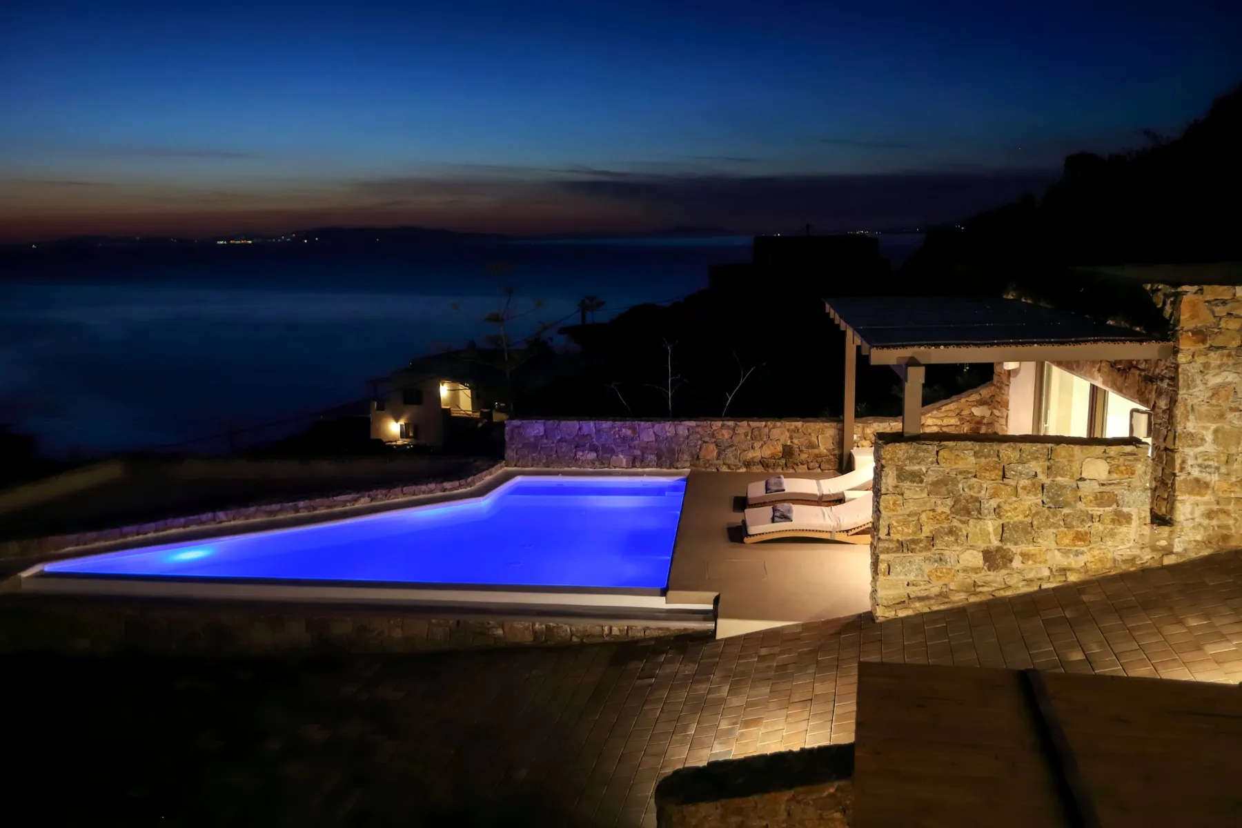An illuminated swimming pool and seating area overlook the water at dusk.