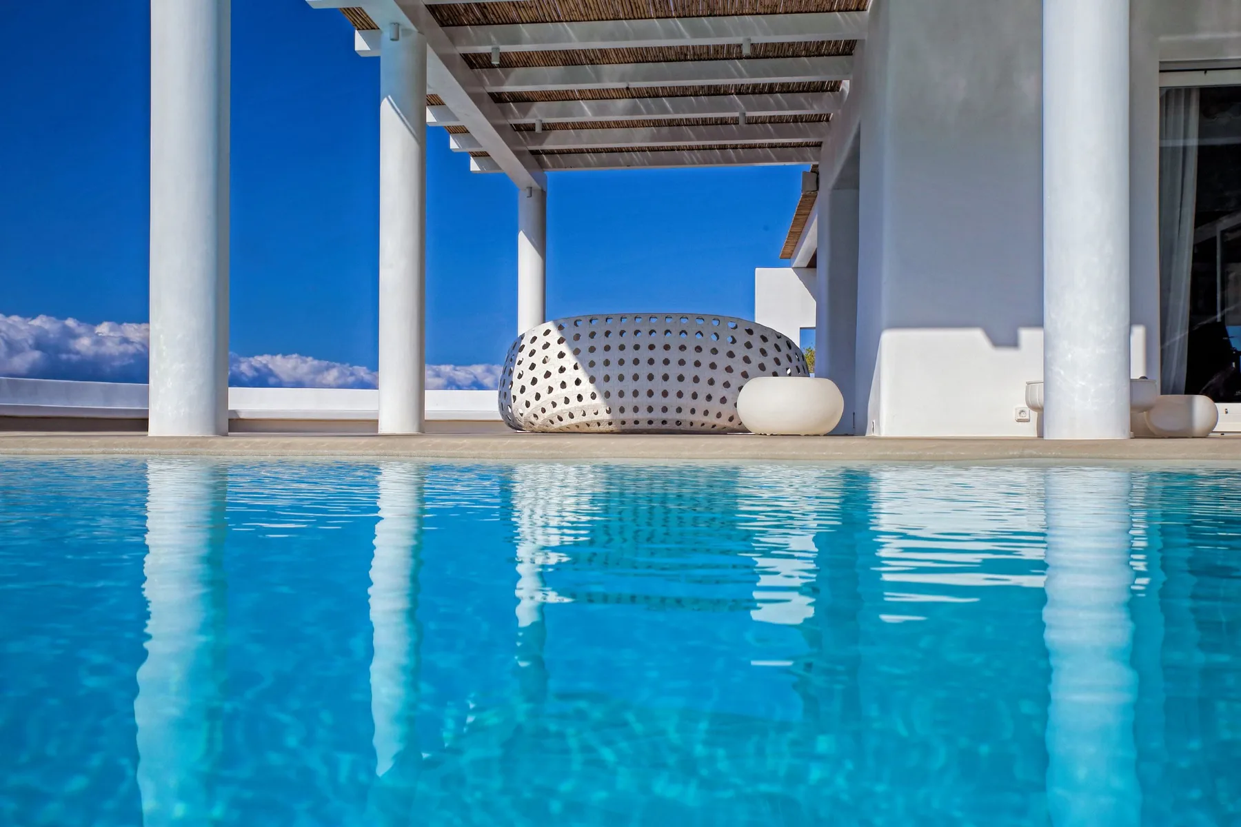 Una piscina al aire libre junto a un edificio blanco, con una zona de estar a la sombra.