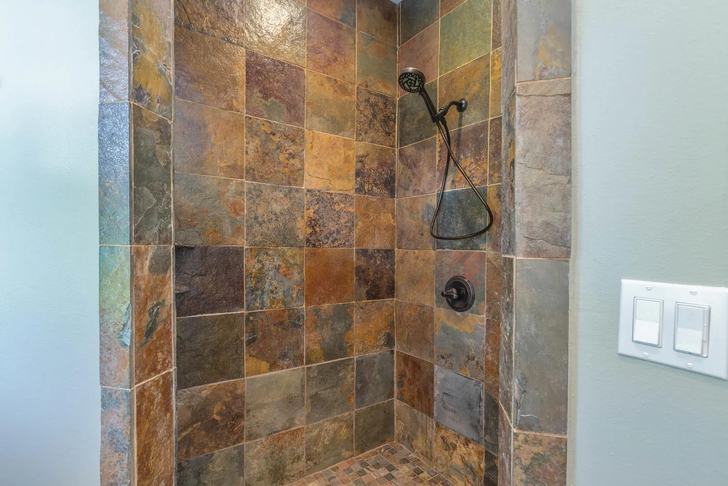 Una ducha con azulejos, cabezal de mano y controles en la pared.
