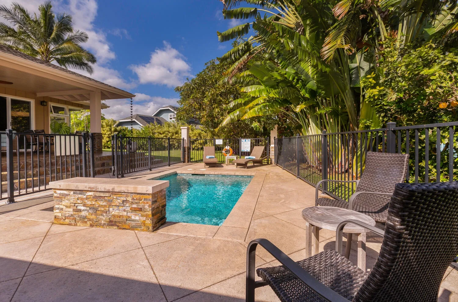 Una piscina exterior está junto a un patio pavimentado con asientos y plantas tropicales.