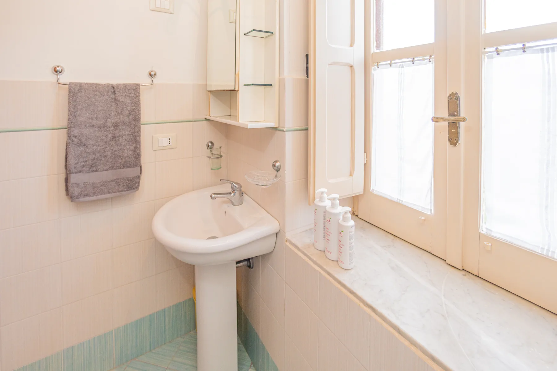 A bathroom sink sits beside a bright window, with a towel hanging on the wall.