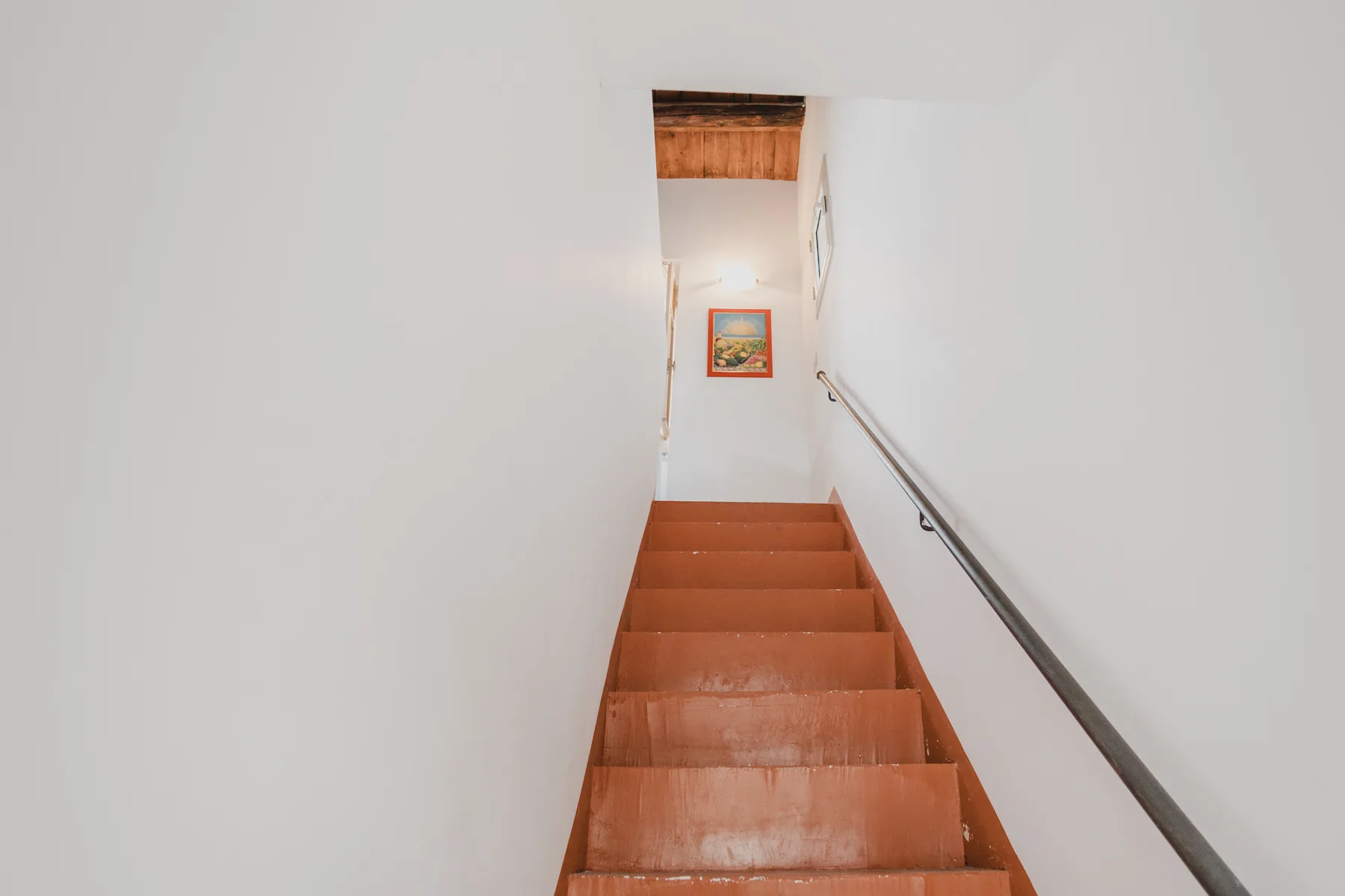 A wooden staircase rises toward a small framed picture on a white wall.