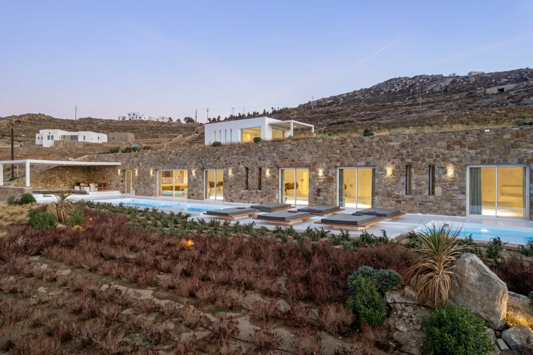 A modern stone building faces a long swimming pool in a dry hillside landscape.