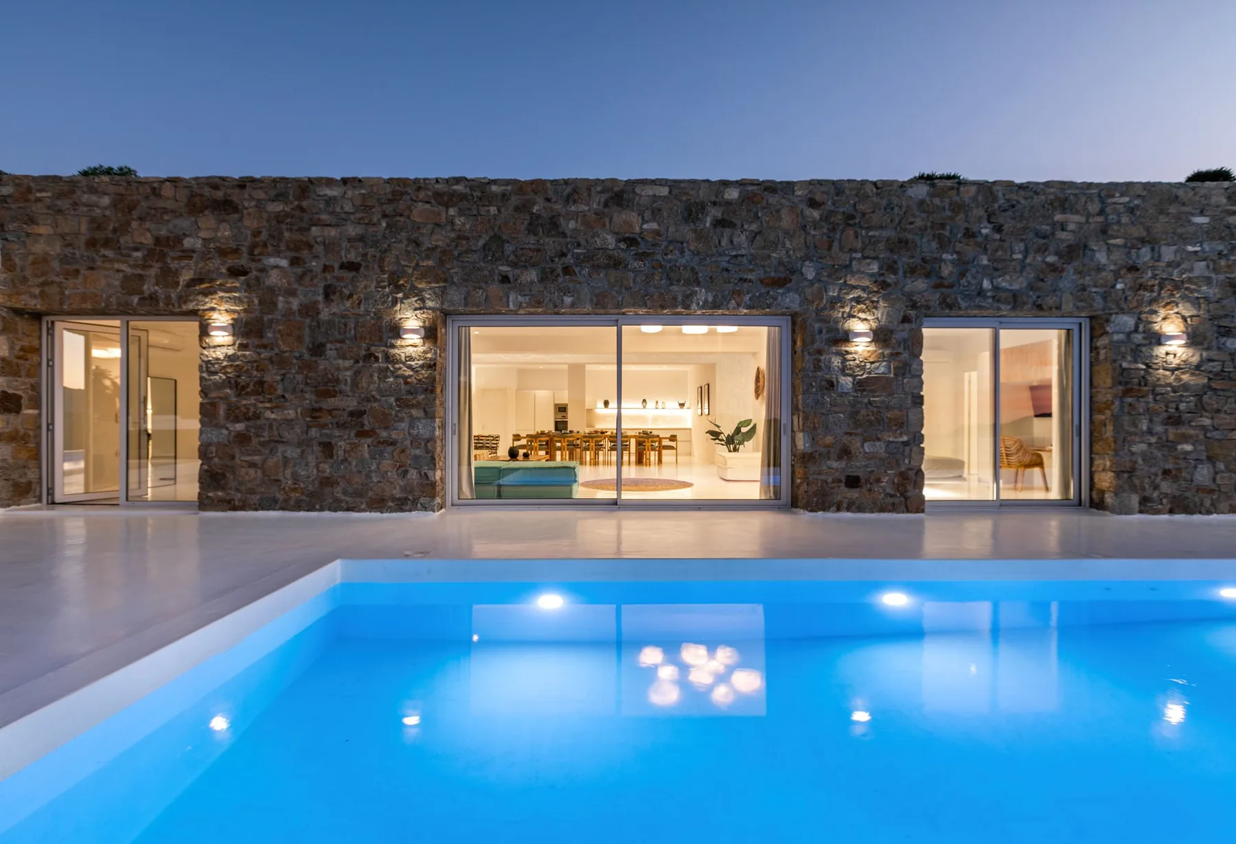 A swimming pool in front of a stone house illuminated at dusk.