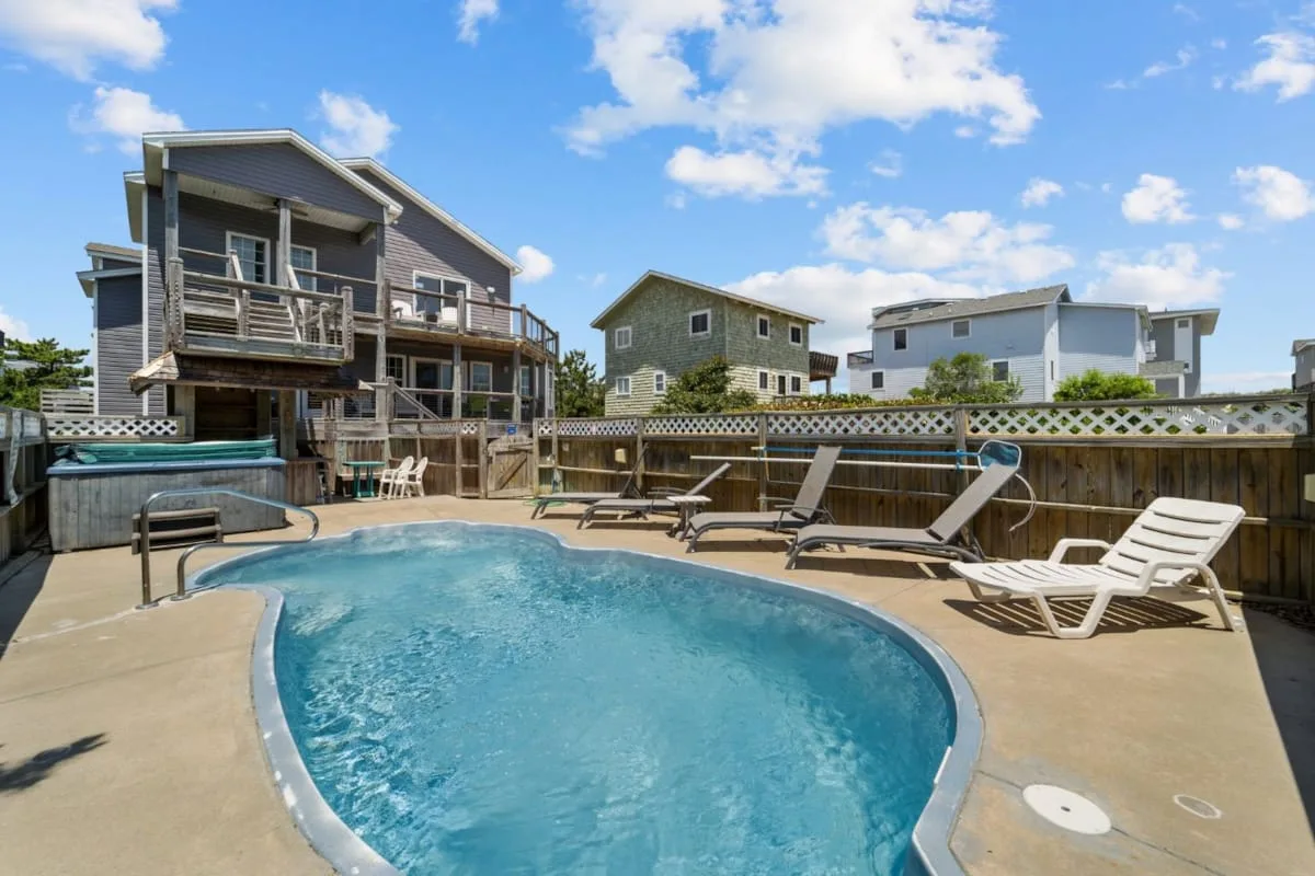 An outdoor pool sits beside a wooden deck with lounge seating and nearby homes.