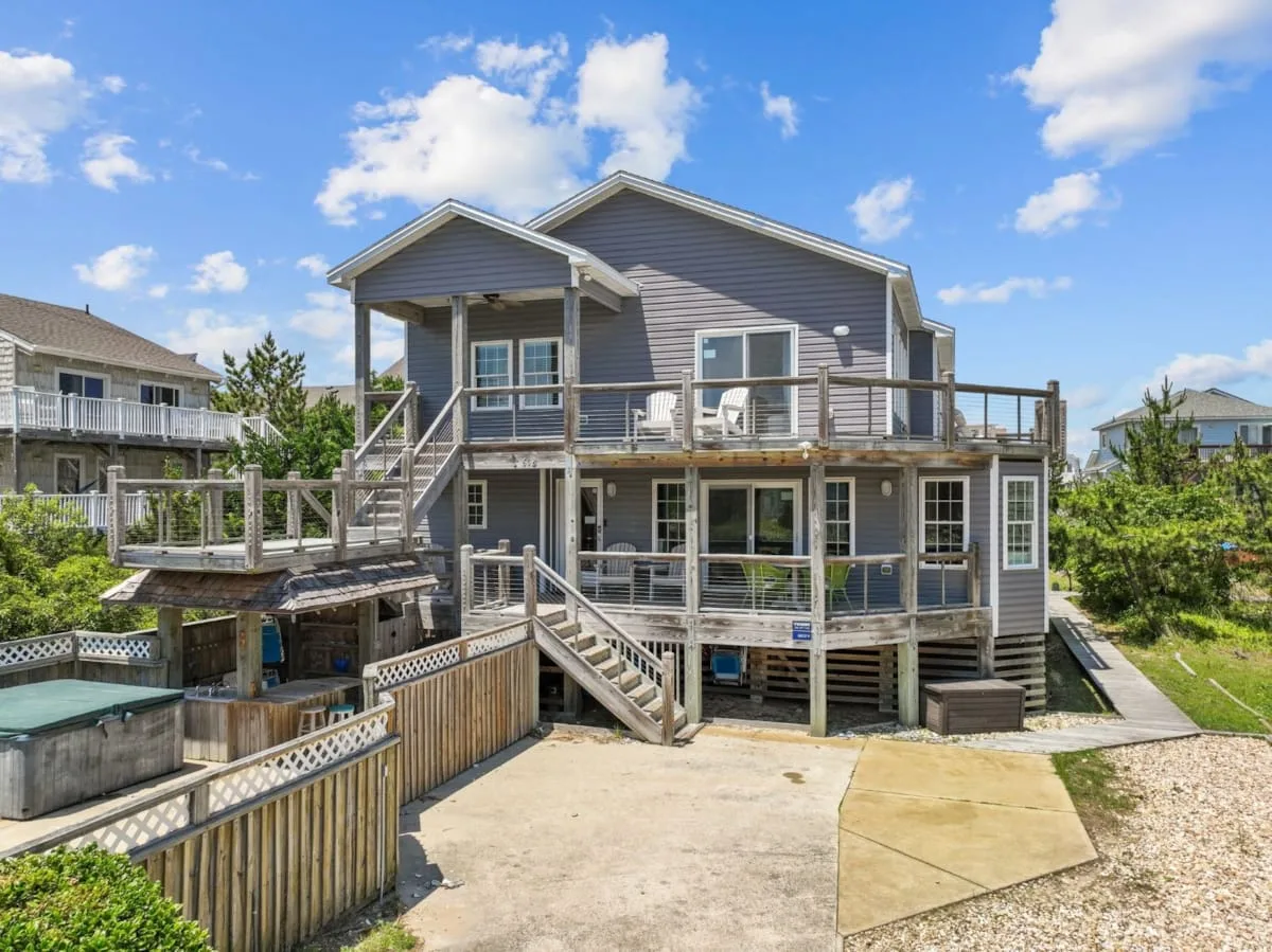 A raised gray house with wooden decks and exterior stairways under a blue sky.