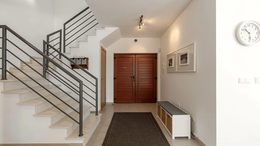 Un recibidor luminoso con una escalera, barandilla oscura y puertas dobles de madera.