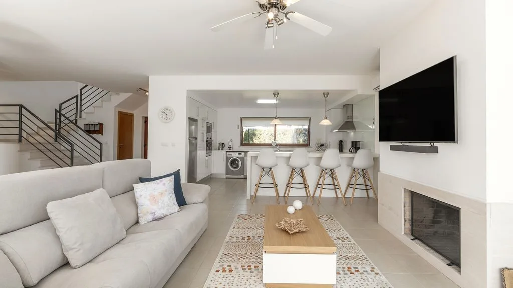 Una luminosa sala de estar se abre a una cocina, con asientos y un televisor en la pared.