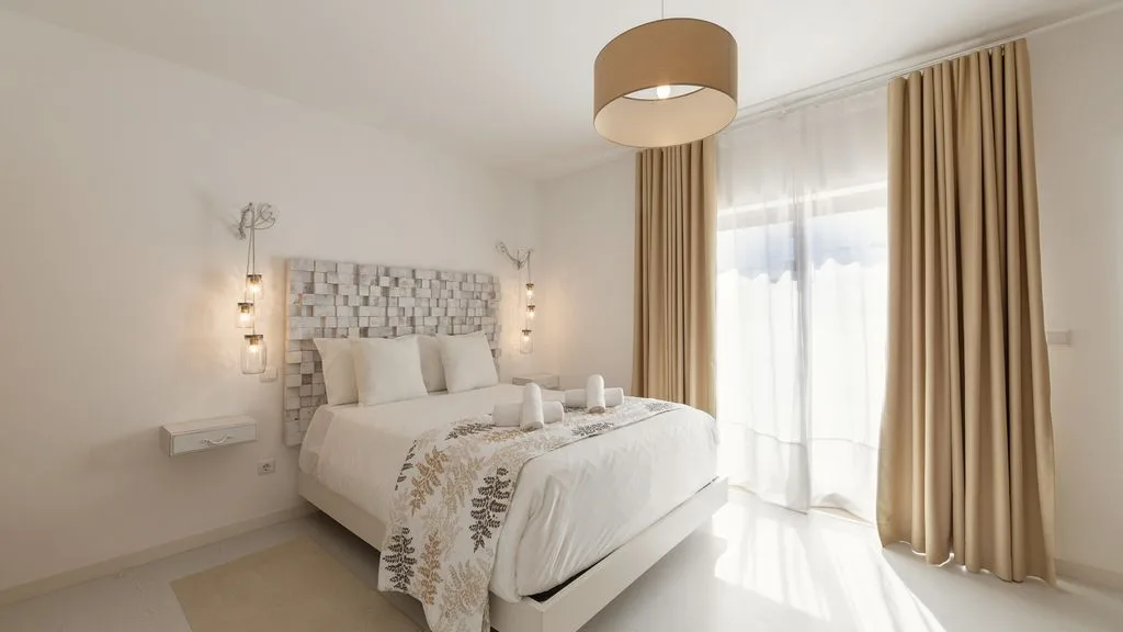 Un dormitorio luminoso con una cama bien hecha junto a altas ventanas con cortinas.
