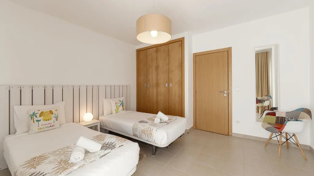 Un dormitorio luminoso con dos camas, un armario de madera y una pequeña zona de estar.