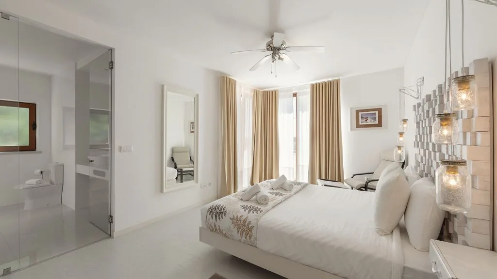 Un dormitorio luminoso con una cama blanca, luces junto a la cama y ventanas altas con cortinas.