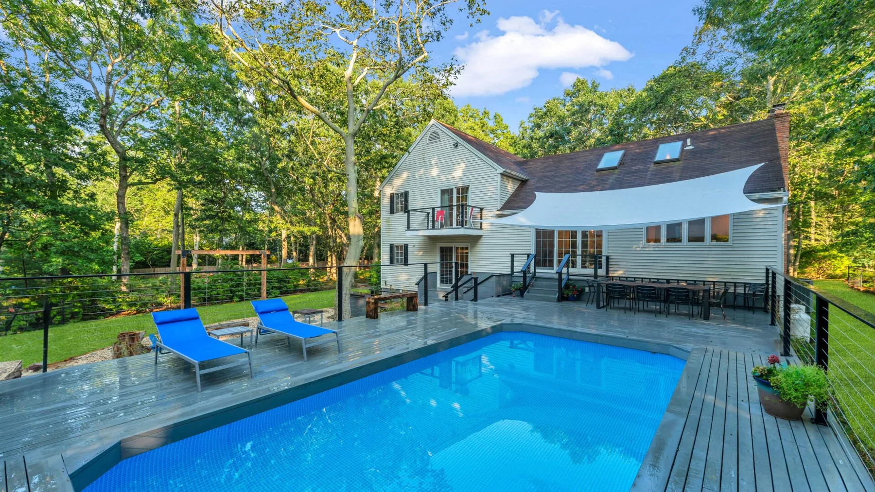A backyard pool beside a house, with deck seating and trees around the yard.