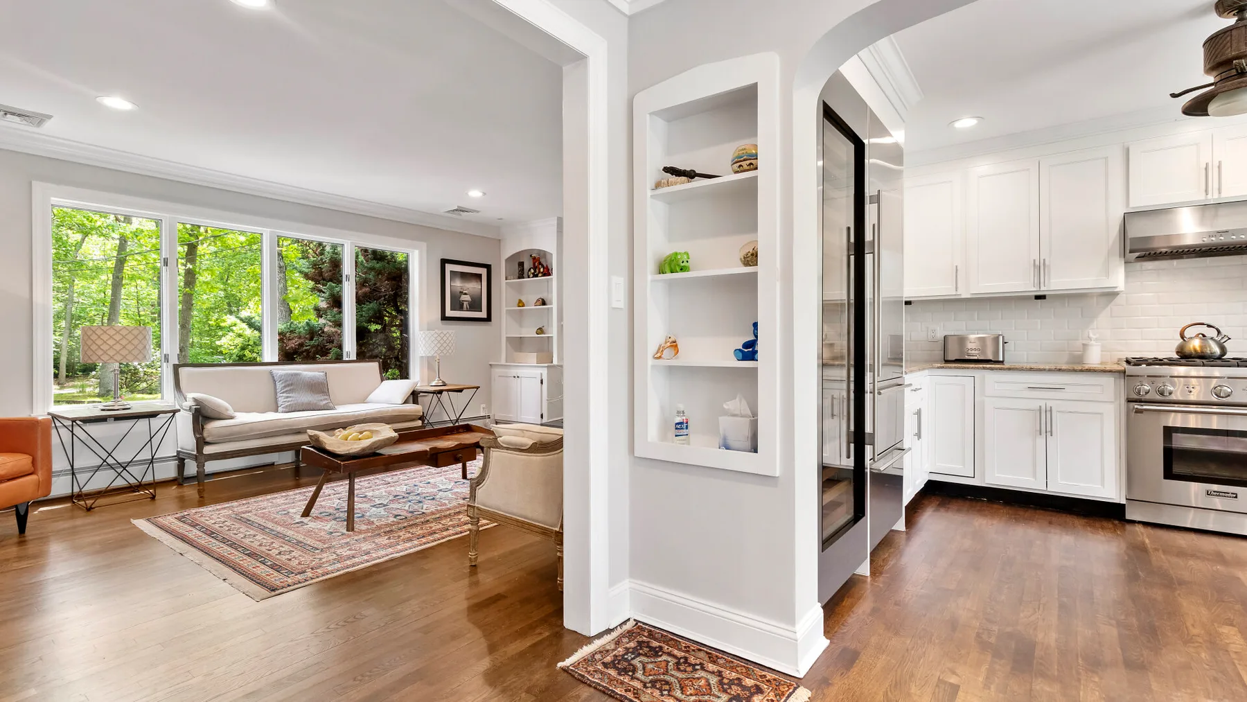 A bright living room opens onto a white kitchen, with large windows and wooden floors.