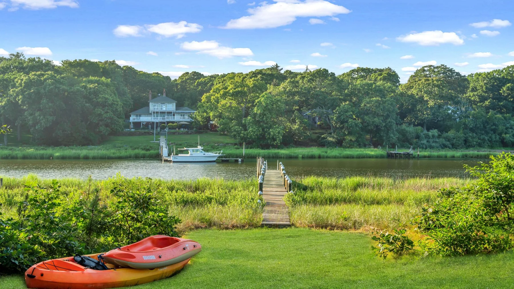 A grassy waterfront opens onto a wooden dock, with a kayak in the foreground.