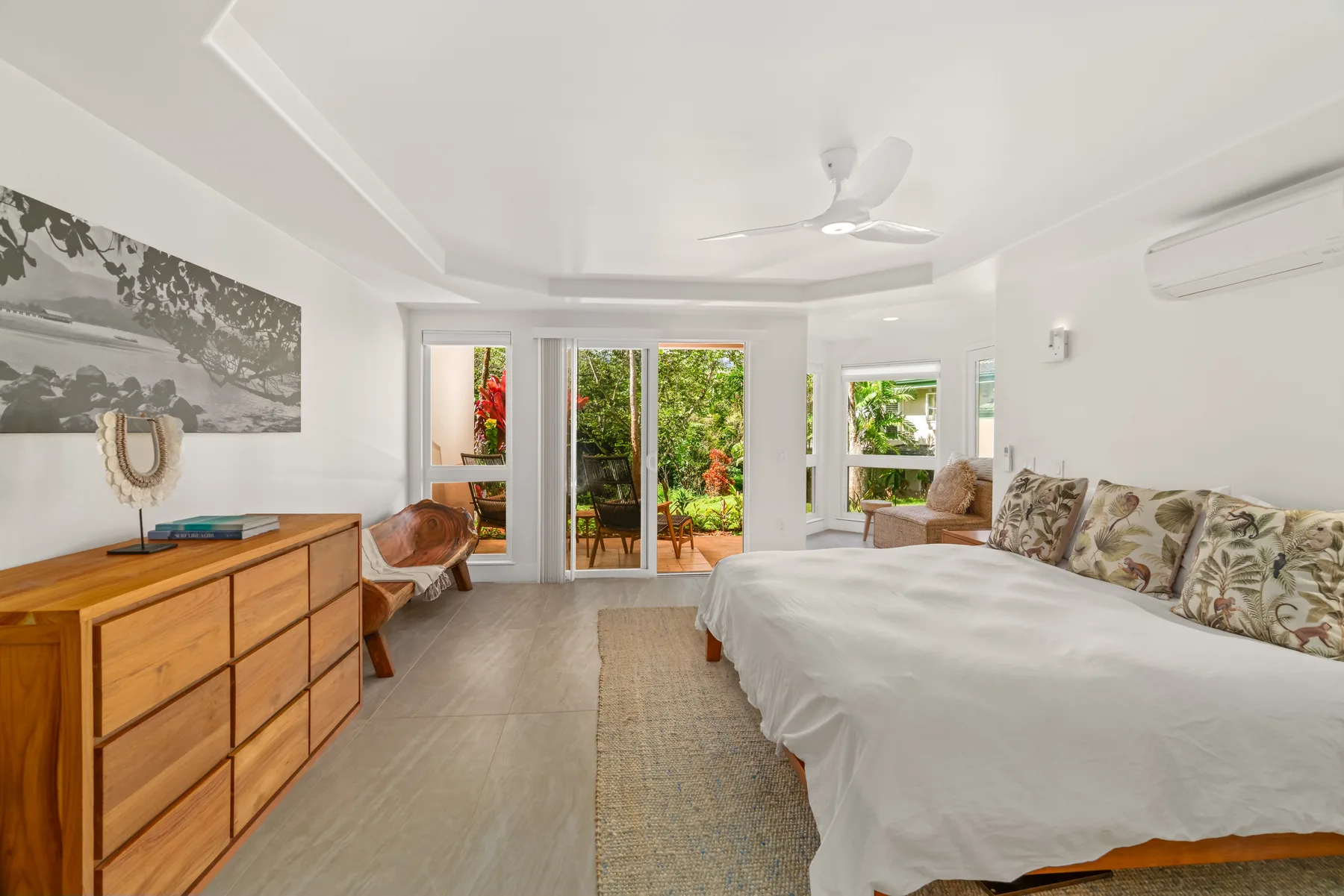 A bright bedroom with a bed, wooden dresser, and windows opening onto greenery.