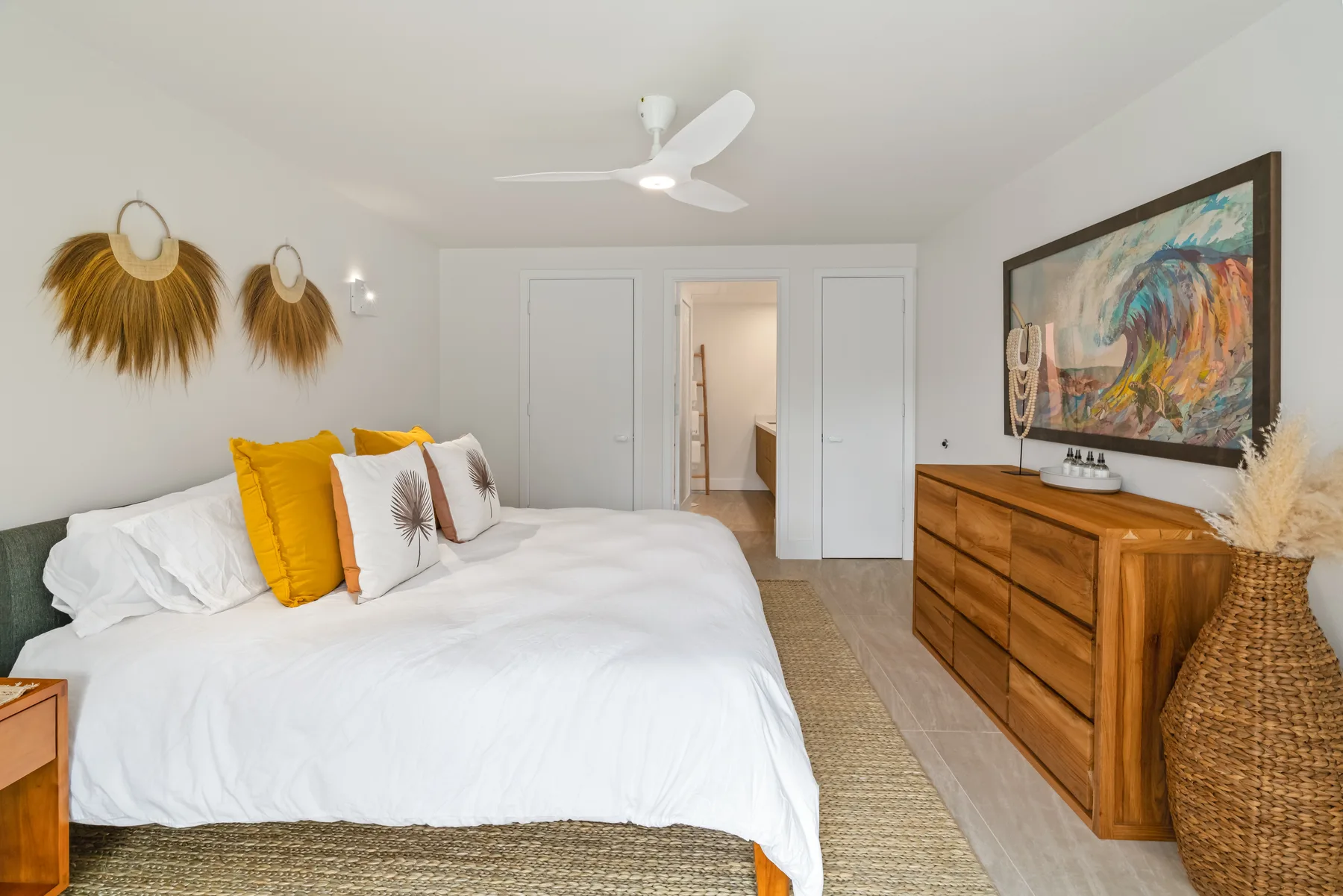A bright bedroom with a white bed and wooden dresser beneath a large painting.