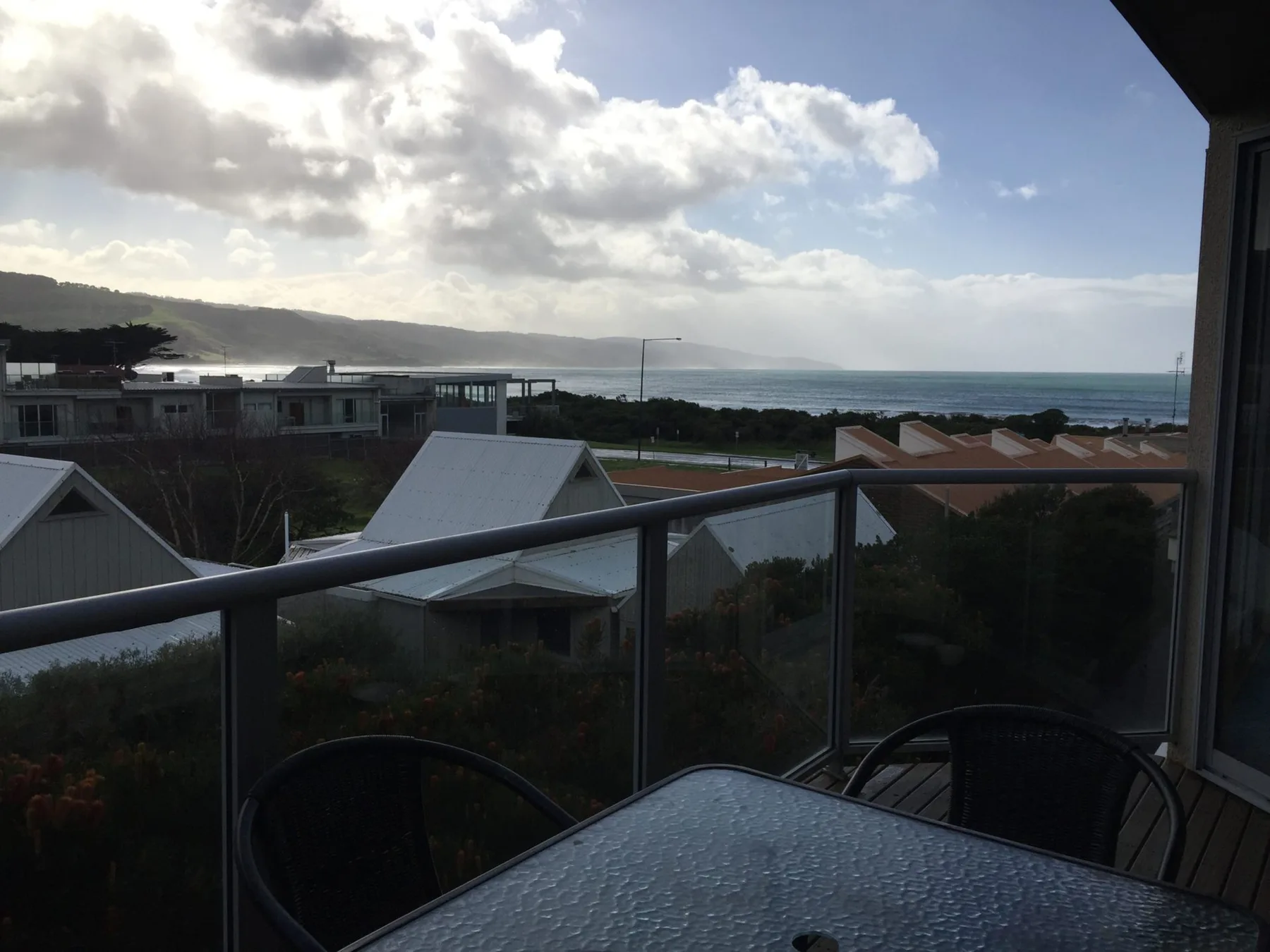 A balcony overlooks nearby rooftops and the ocean beneath a cloudy sky.