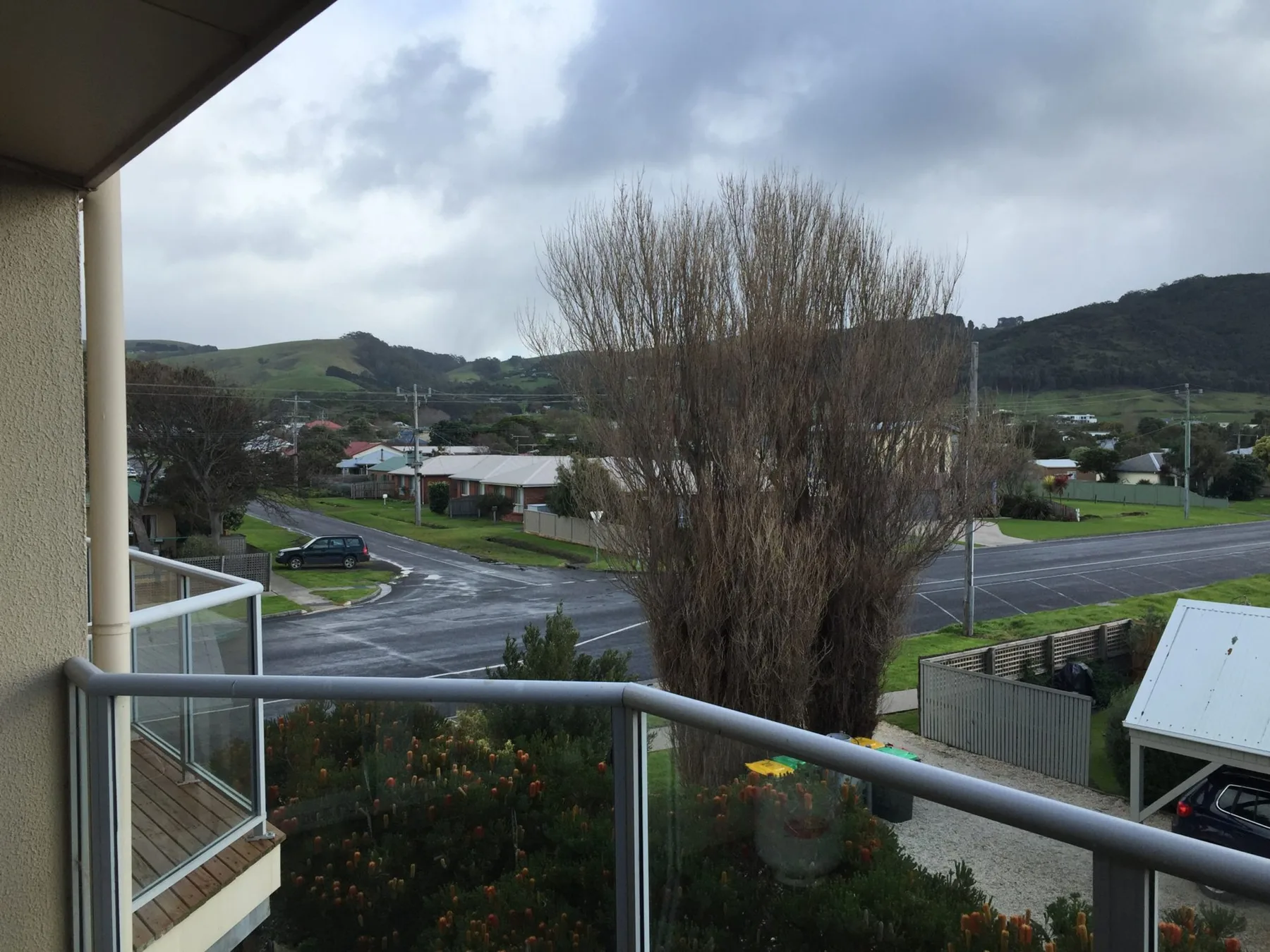 A balcony overlooks a residential street and green hills under a cloudy sky.