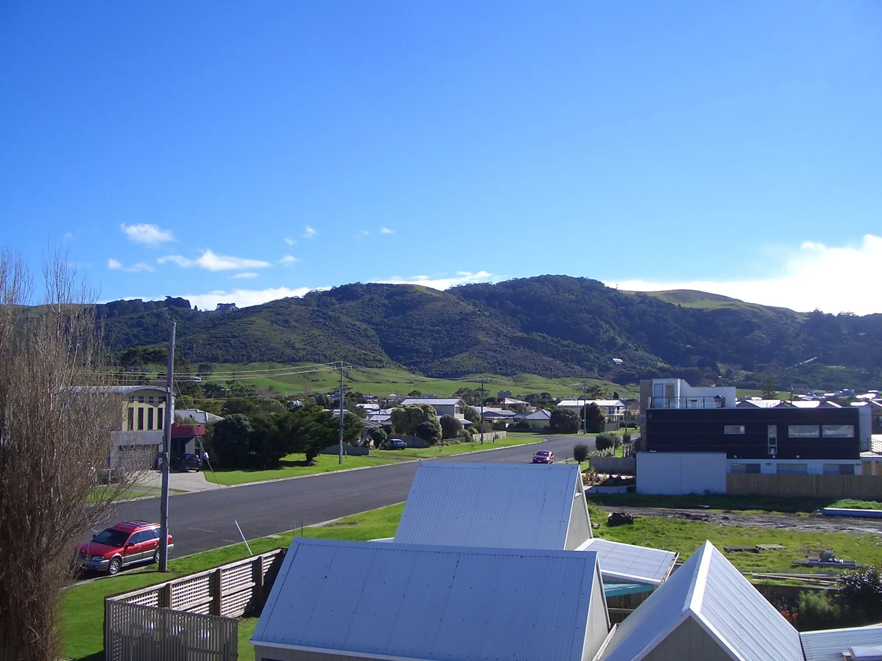 A roadside scene with low buildings, greenery, and hills under a clear sky.