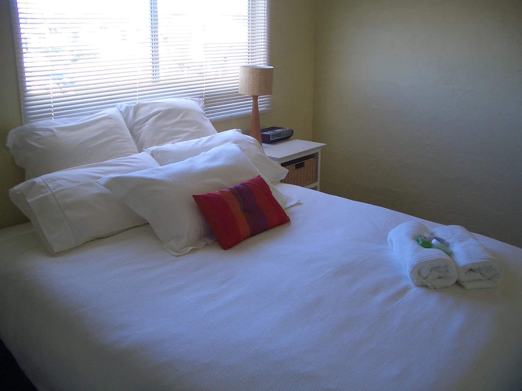 A bedroom with white bedding, a colorful accent pillow, and a window with blinds.