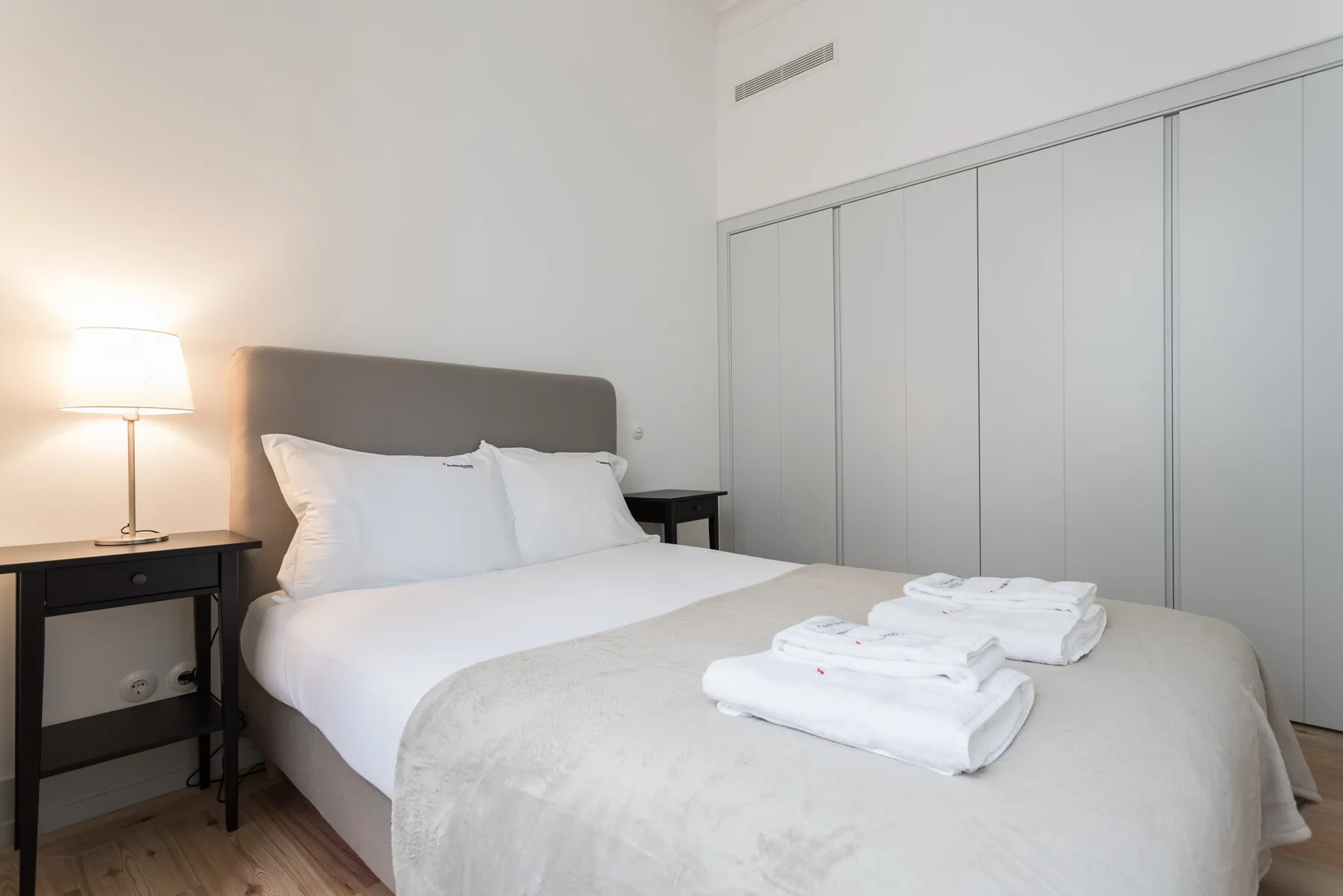 A bedroom with a neatly made bed, folded towels, and a bedside lamp.