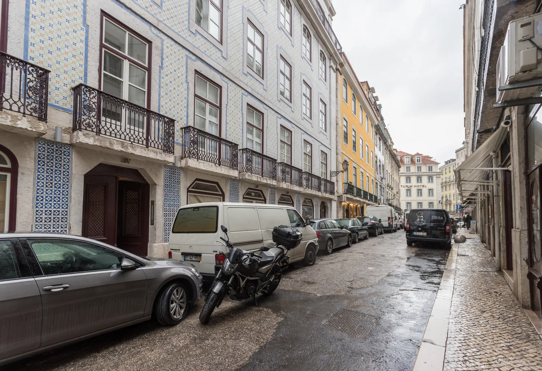A narrow city street lined with tiled buildings and parked vehicles.