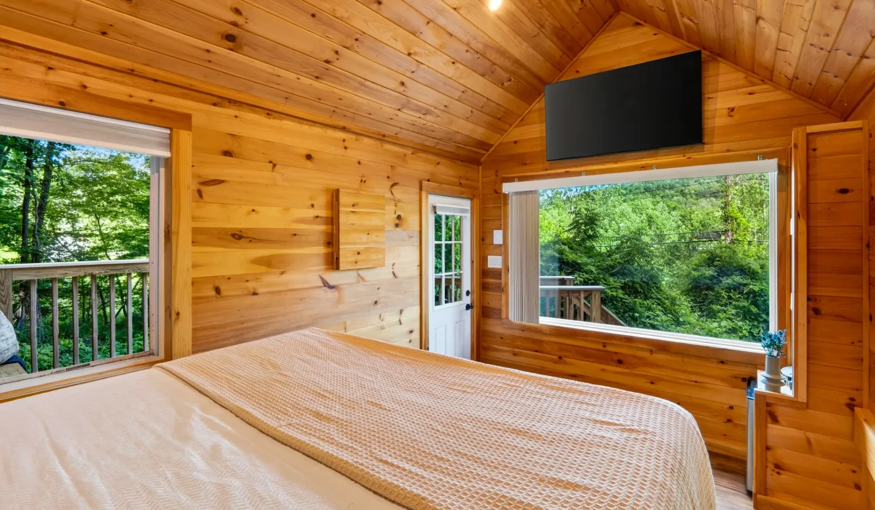 Un dormitorio revestido de madera con una cama y grandes ventanas con vistas a árboles verdes.
