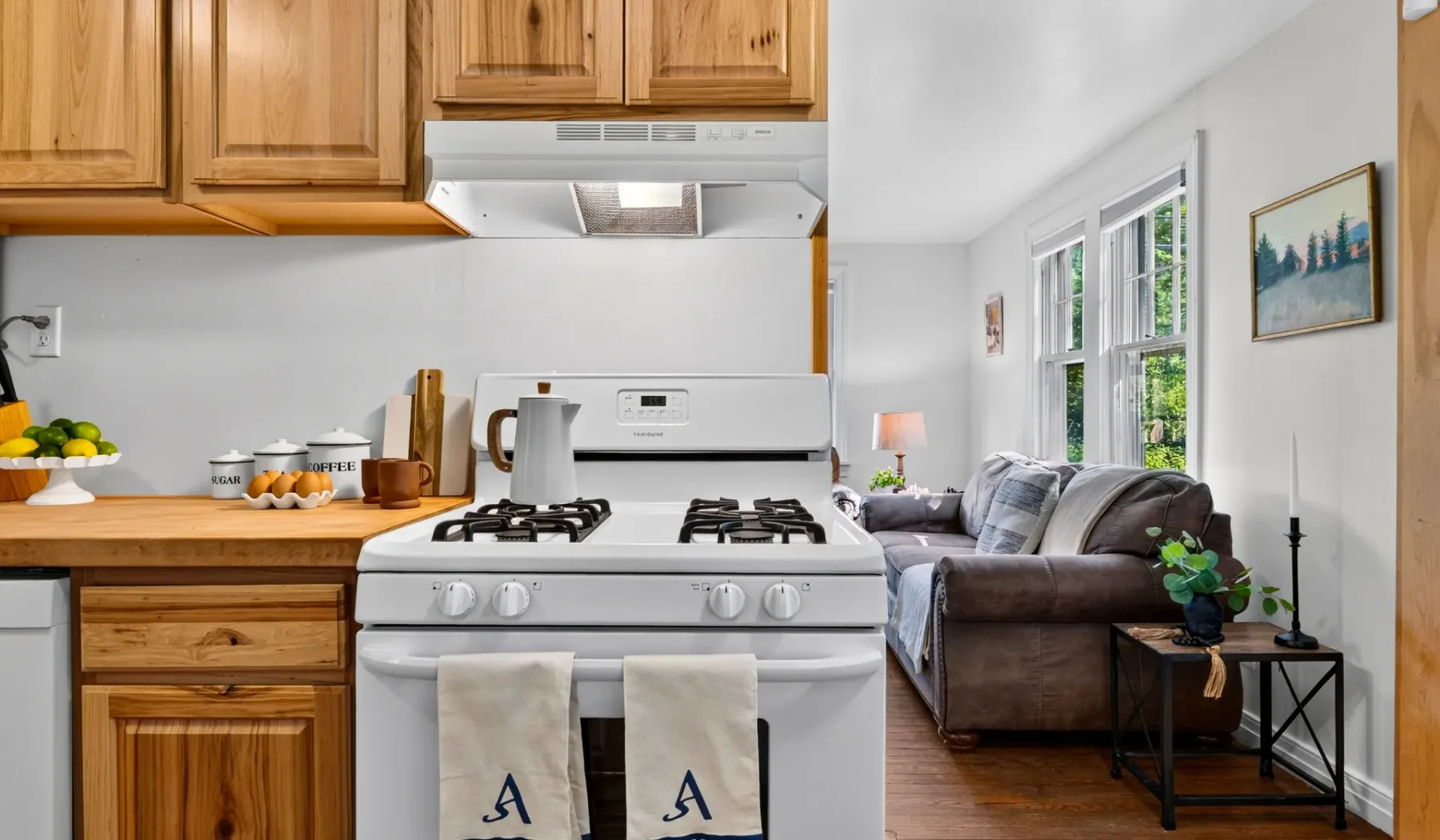 Una cocina se abre a una sala de estar con un sofá junto a las ventanas.