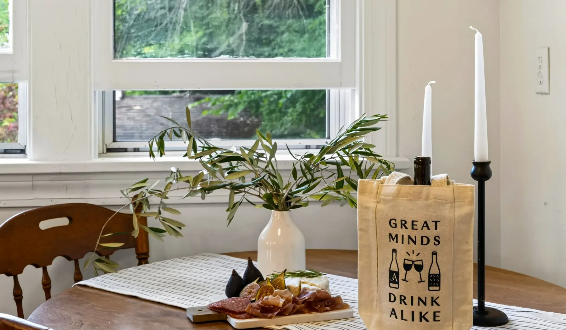 Una mesa de comedor junto a grandes ventanas tiene un jarrón, velas y una tabla para servir.
