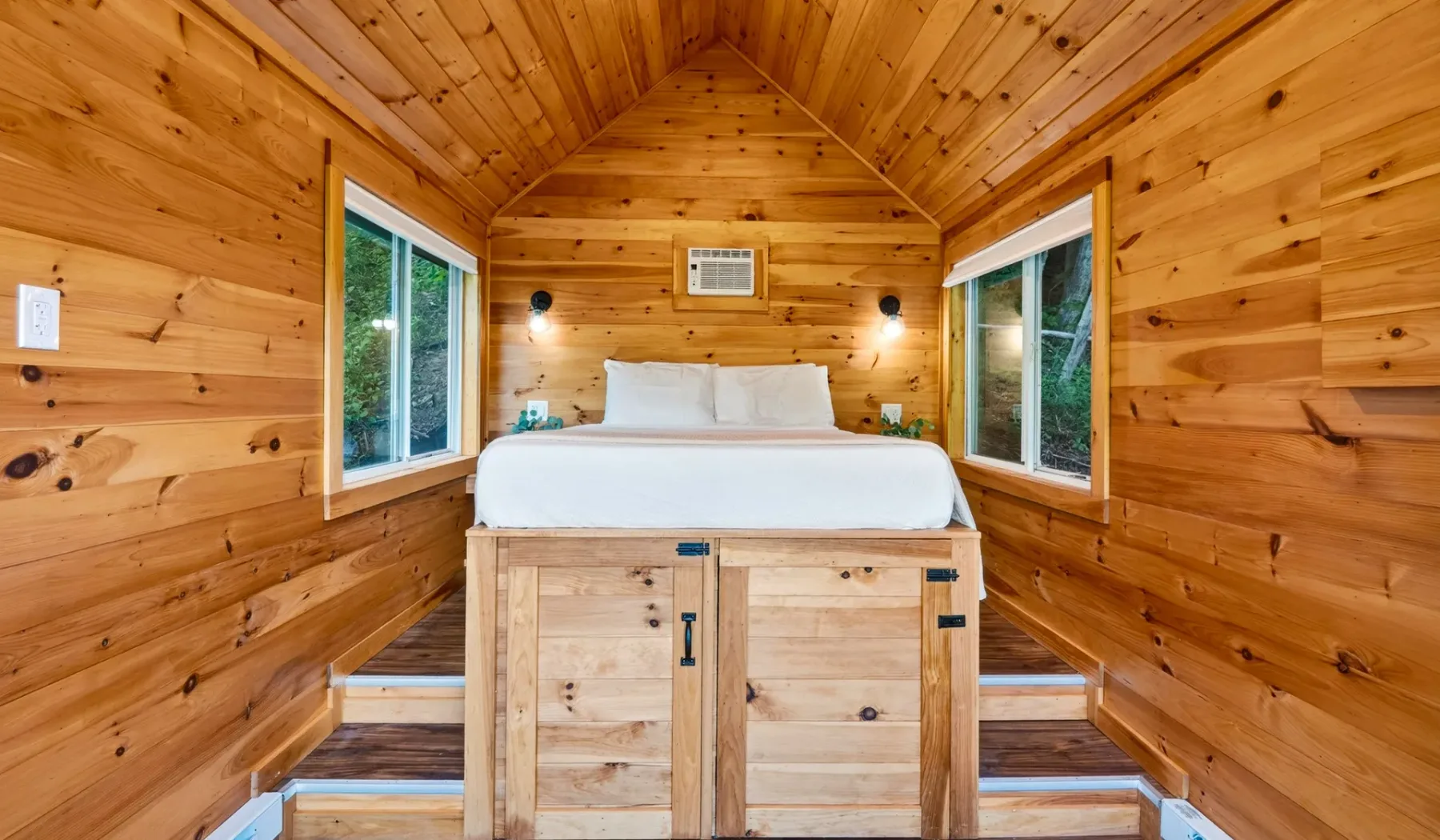 Un dormitorio con una cama entre dos ventanas en una habitación revestida de madera.
