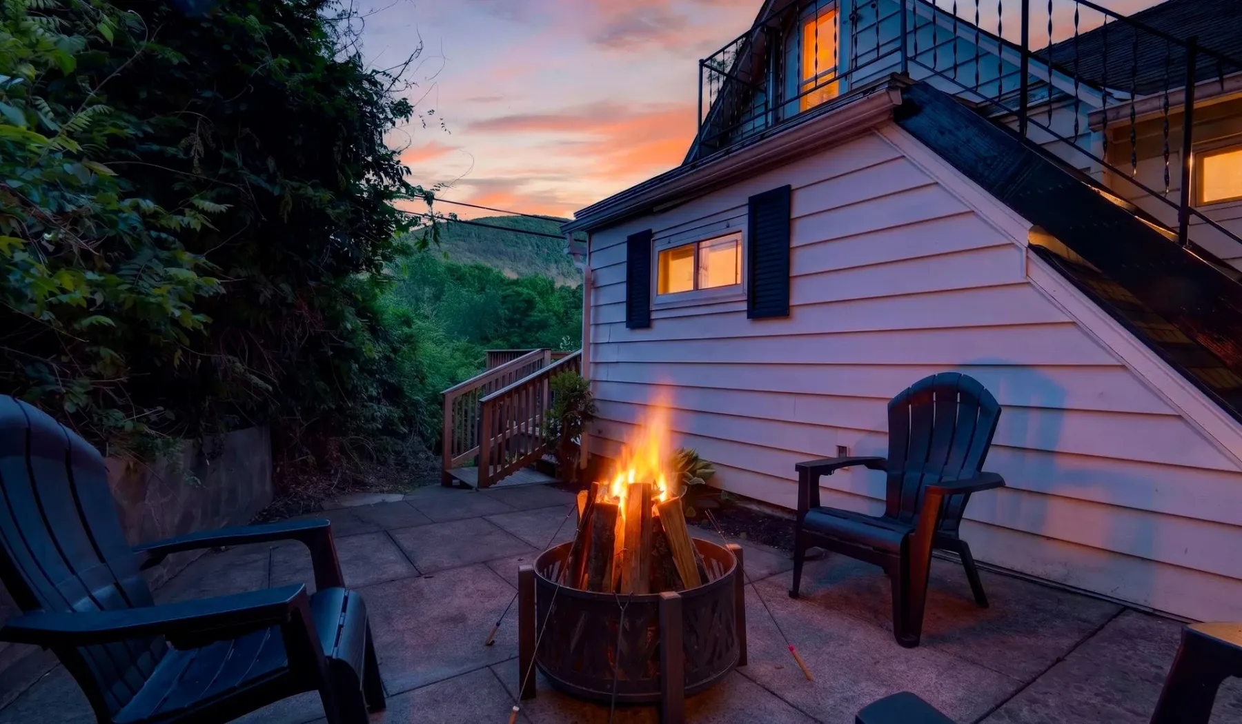 Un patio exterior con asientos y un brasero encendido junto a una casa al atardecer.