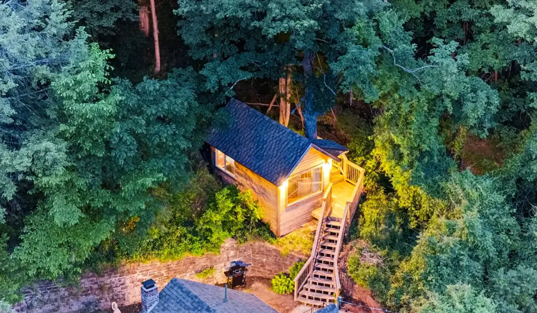 Una pequeña cabaña de madera está entre árboles, iluminada por luces cálidas.