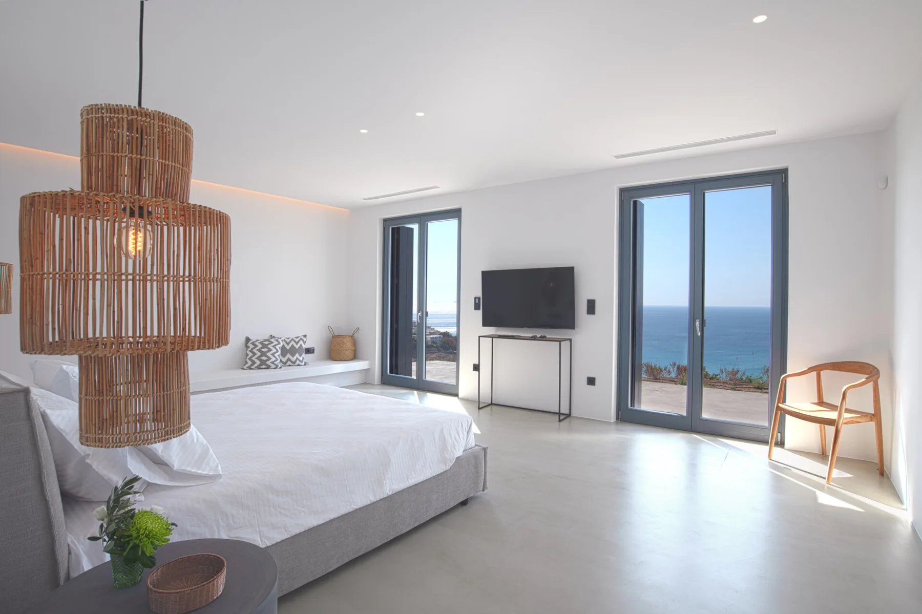 A bright bedroom with a bed, television, and glass doors overlooking the sea.