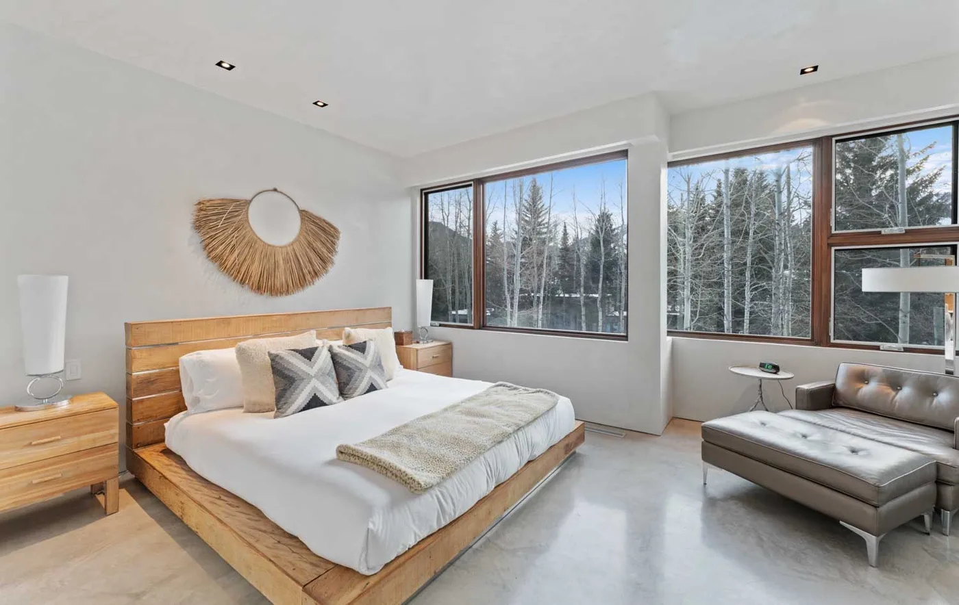 A bright bedroom with a wooden bed and large windows overlooking trees.