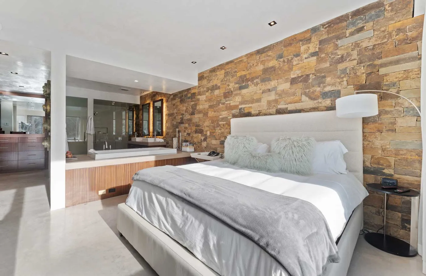 A bedroom with a bed against a textured stone wall and a bathroom area beyond.