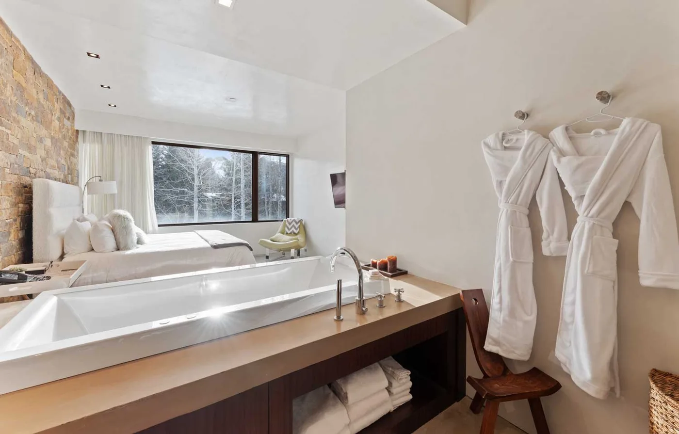 A bright bathroom features a wide tub, a large window, and hanging robes.