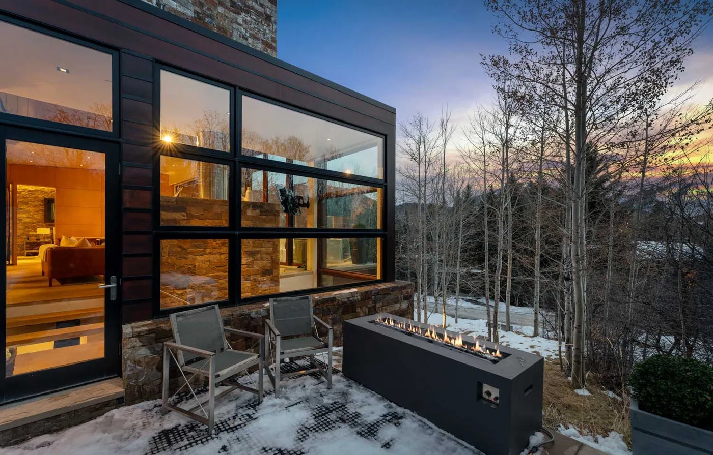 A modern home opens onto a snowy patio with outdoor seating and a long fire feature.