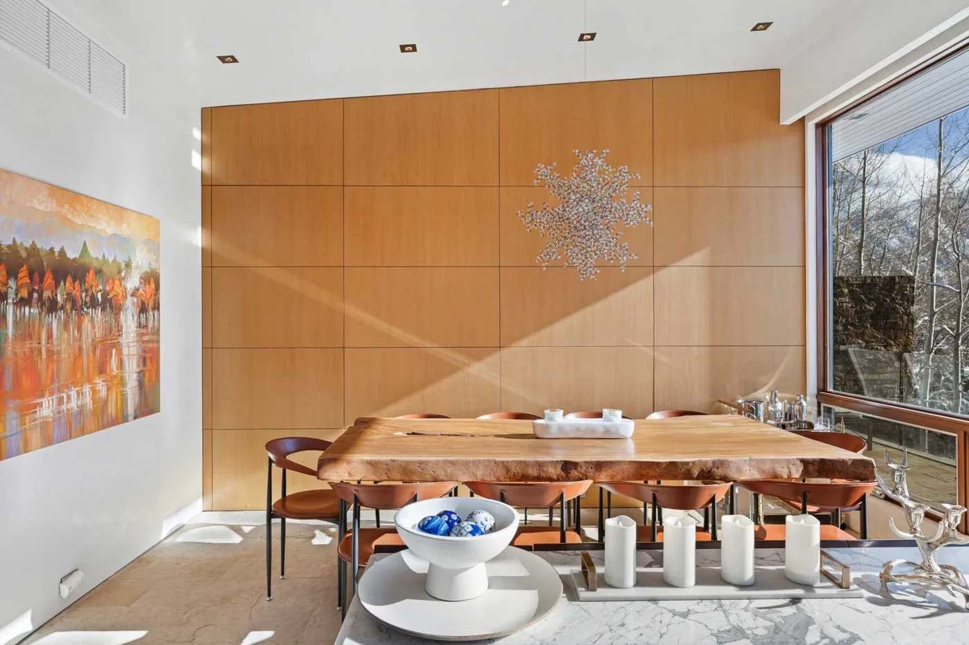 A dining area with a long wooden table, sculptural wall art, and large windows.