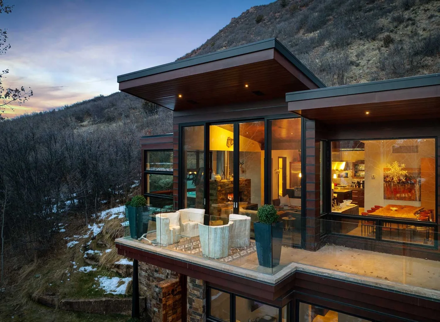 A modern hillside house with large glass windows and a furnished terrace at dusk.
