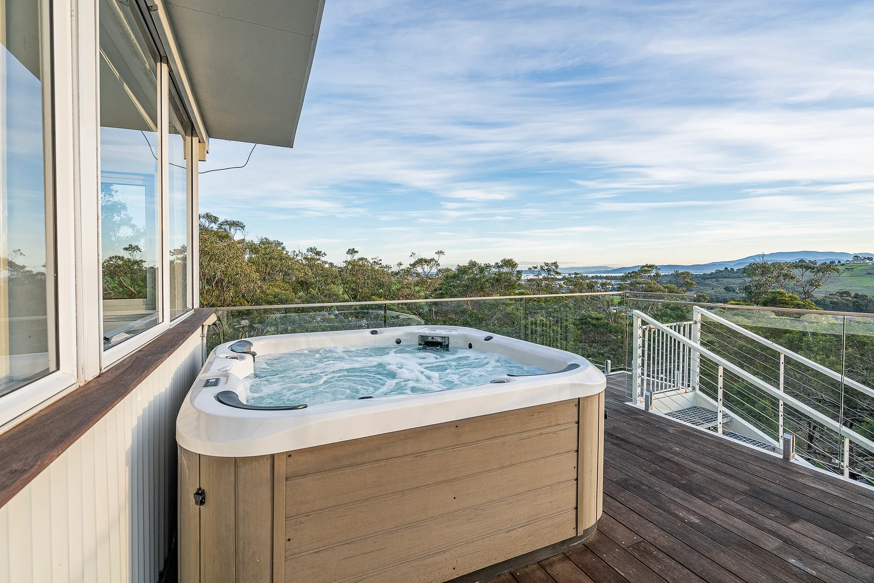 An outdoor spa sits on a deck beside a glass railing, with trees and distant hills beyond.