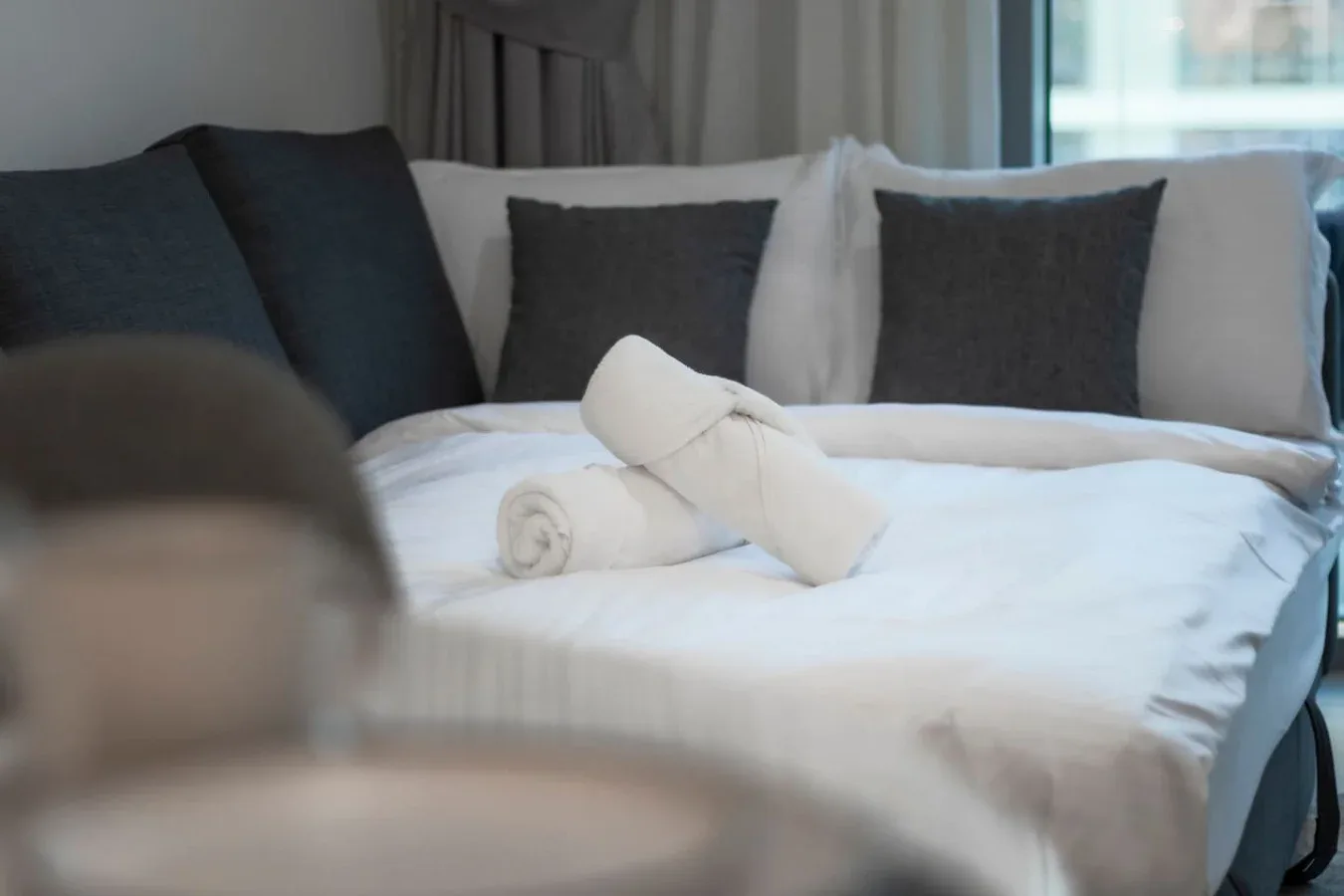 Una cama con cojines oscuros y toallas blancas dobladas está junto a una ventana.