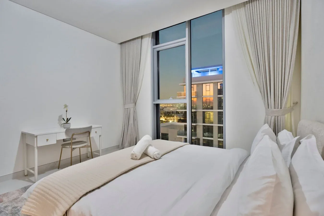 Un dormitorio con ropa de cama blanca, un escritorio y ventanas altas con vistas a edificios cercanos.