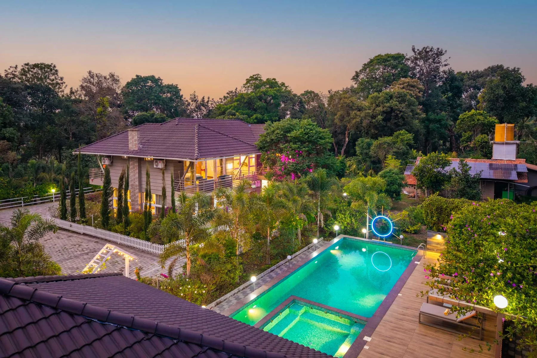 Una piscina exterior ajardinada está junto a un edificio iluminado, rodeada de árboles.